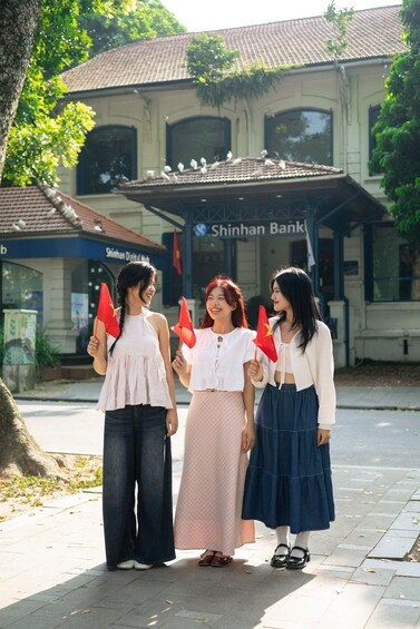 Three women holding red flags in front of a building with a tiled roof and a sign that reads 'Shinhan Bank.' 3