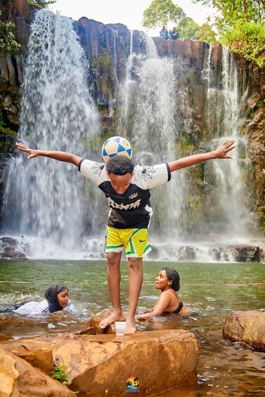 A person balancing a soccer ball on their head, standing on rocks by a waterfall. 3
