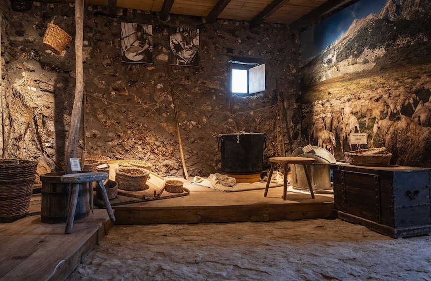 A rustic interior with stone walls, a wooden beam ceiling, a window, and various traditional items like baskets and a large pot. 2