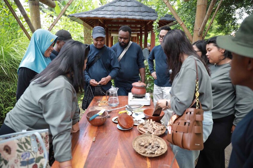 A group of people around a wooden table with cooking utensils and food items, in a natural, outdoor setting with trees and a traditional structure in the background. 5