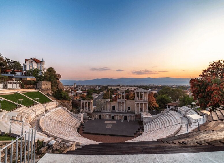 An ancient amphitheater with stone seating and ruins, surrounded by modern buildings and a clear sky. 1