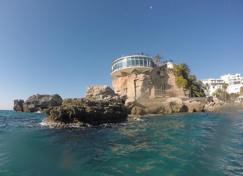 A coastal rock formation with a circular building on top, clear blue sea, and white buildings in the background. 1