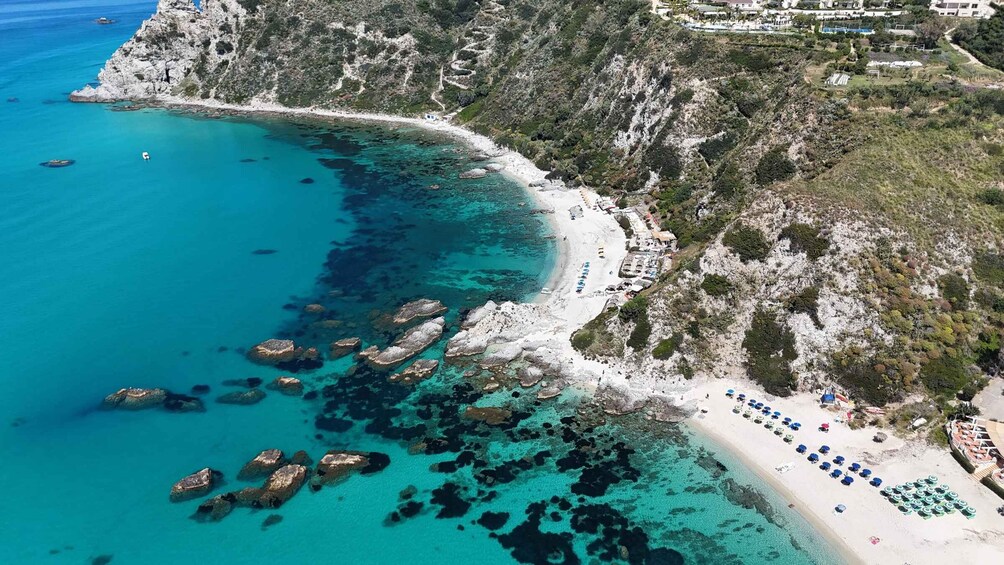 A coastal landscape with clear turquoise waters, rocky outcrops, and a sandy beach with umbrellas. 3