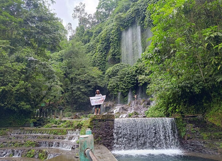 A waterfall surrounded by lush greenery, a person standing on a rock with a sign, and a small stream flowing through the scene. 2