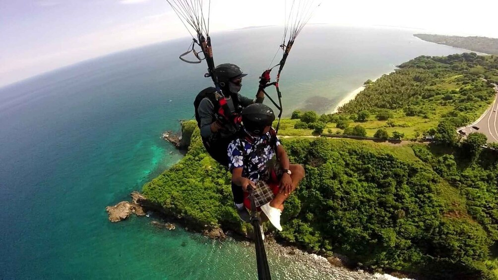 A person paragliding over a coastal area with clear turquoise waters and lush green hills. 1