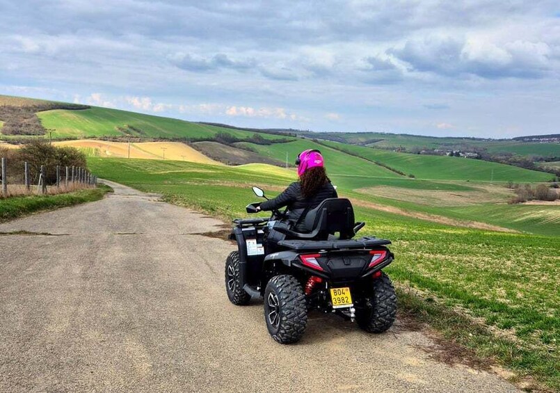 A person on a black ATV with a pink helmet, driving on a rural road surrounded by green fields and hills. 4
