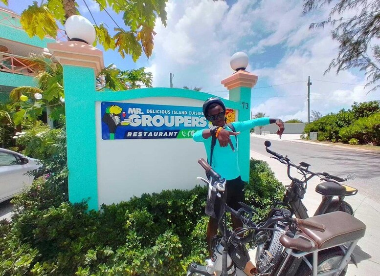 A person standing next to a sign for 'Mr. Delicious Island Cuisine Groupers Restaurant' with a motorcycle parked nearby. 1