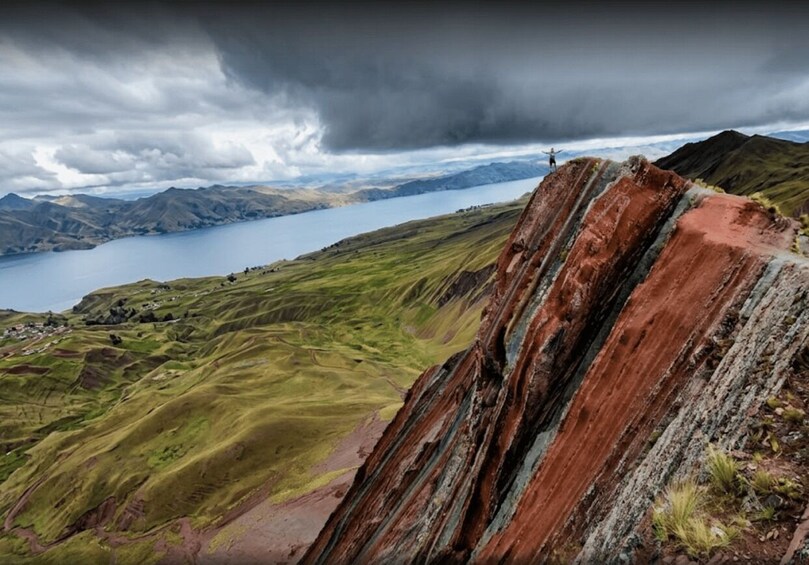 A rugged cliff with a person standing at the edge, overlooking a valley with a lake and distant mountains under a cloudy sky. 2