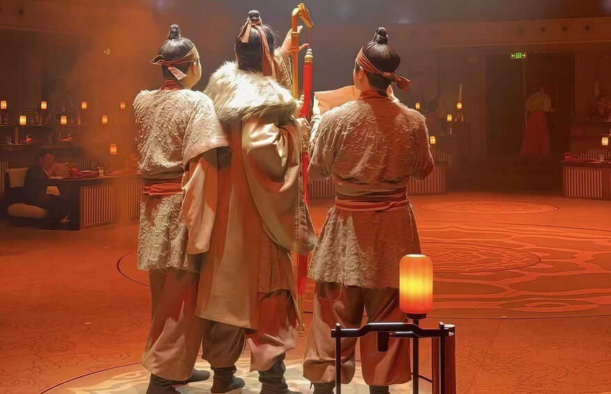 A group of people in traditional attire, standing in a dimly lit room with a patterned carpet and a table with a lamp. 4