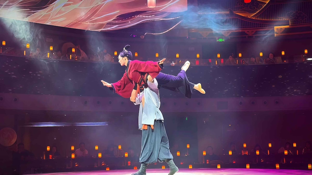 A performer in traditional attire executing a lift with a partner on a stage with an audience. 3
