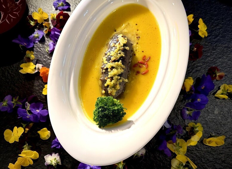 A white bowl with a dark-colored food item, possibly a meat or fish, topped with yellow grains and garnished with red chili flakes. A piece of broccoli is placed beside the bowl. 5