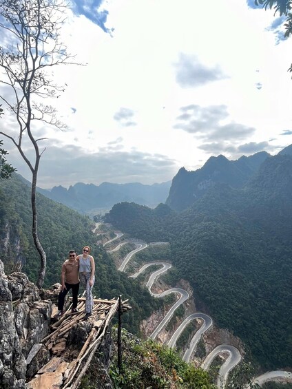 Two people standing on a wooden platform overlooking a winding mountain road. 3