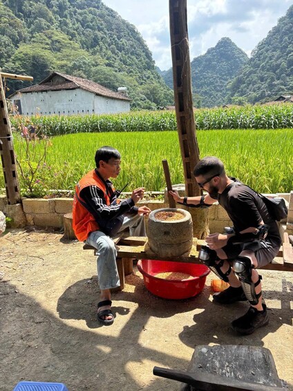 Two individuals grinding grains using a traditional stone mill, with a rural landscape and a house in the background. 1