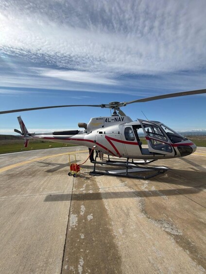 En helikopter med beteckningarna ”4L-NAV” och ”AIRBUS” på sidan, parkerad på en betongyta med en klar himmel i bakgrunden. 2