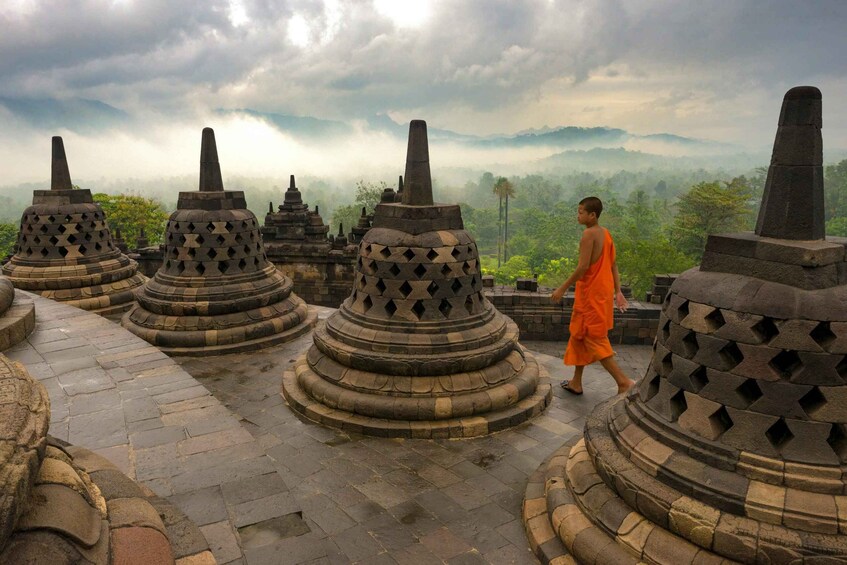 A person in orange robes walking between stone stupa structures with a mountainous landscape in the background. 3