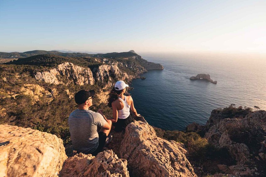 A couple sitting on a rocky outcrop overlooking a coastal landscape with cliffs and a small island in the distance. 1