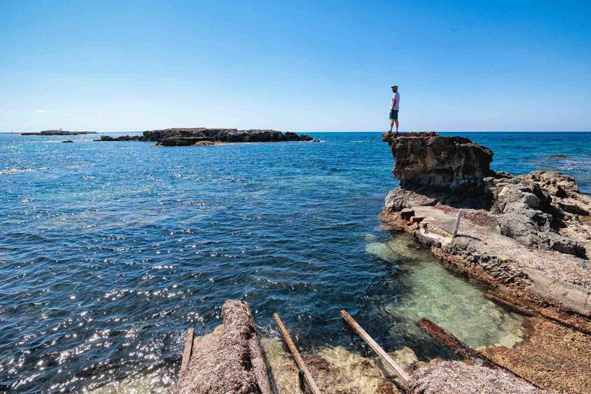 A person standing on a rocky outcrop by the sea, with clear water and distant landforms. 5