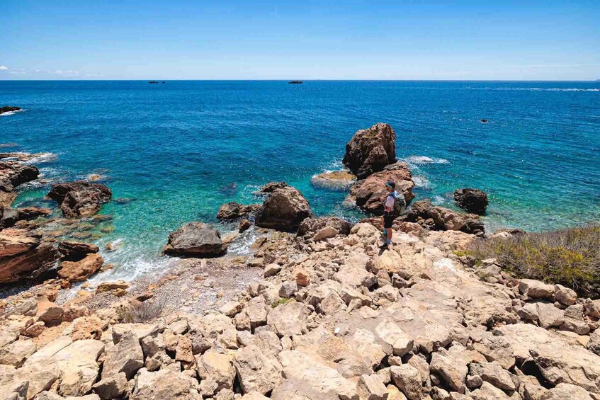 A rocky coastline with clear blue waters and a person standing on the rocks. 1