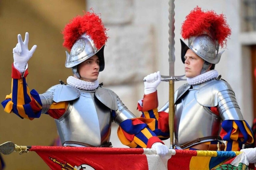 Two individuals in medieval armor with red plumes, holding a flag. 2