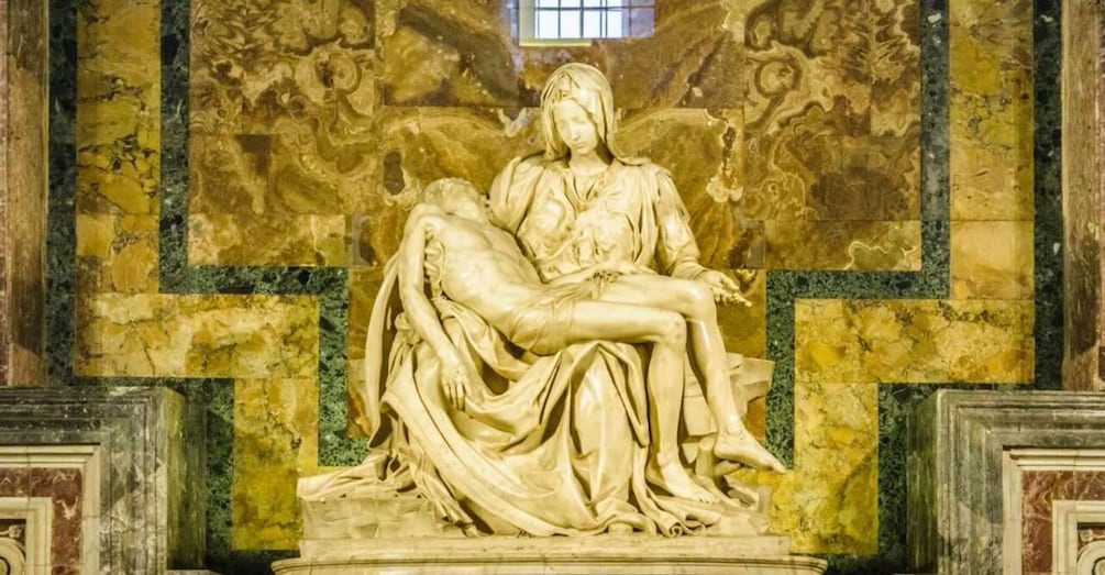 A marble sculpture of a seated figure holding a reclining figure, set against a patterned wall with a window above. 4