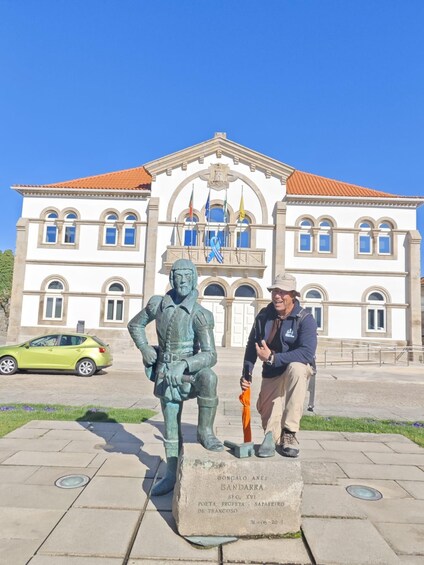 A statue of a historical figure, a man in armor, and a plaque with text in front of a building with a red roof and flags. 5