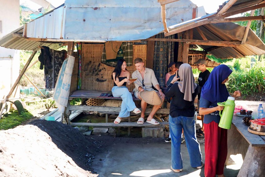 A group of people sitting and standing under a makeshift shelter with a wooden bench and a corrugated metal roof. 2