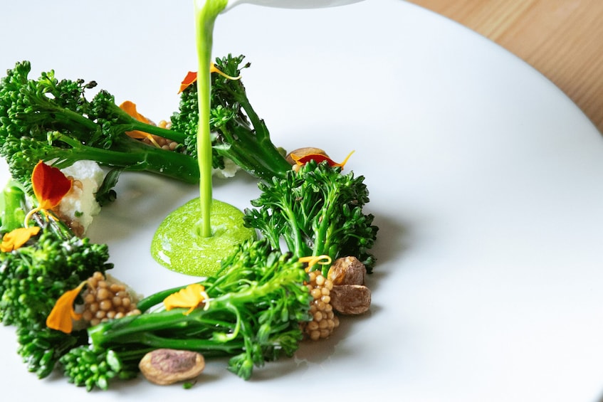 A white plate with a variety of vegetables, including broccoli and carrots, and a green sauce being poured over them. 1