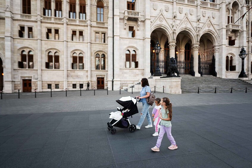 A woman pushing a stroller with two children in front of a historic building with arched windows and a statue. 3