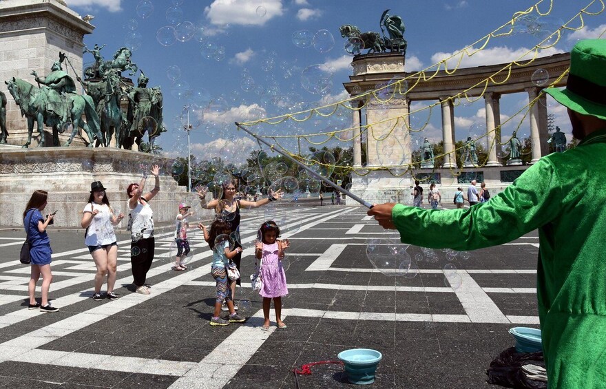 A group of people playing with bubbles in a public square with statues and a fountain. 5