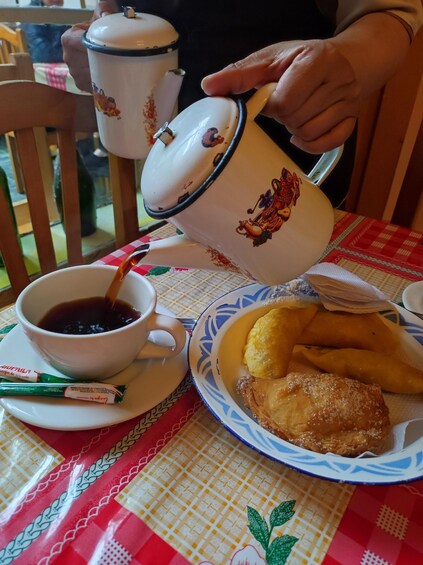 A person pouring coffee into a cup, a plate with pastries, and a teapot on a table with a checkered tablecloth. 2