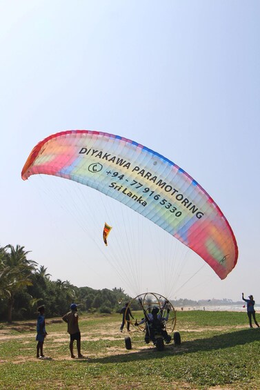 A paraglider with a colorful canopy, a person standing on the ground, and another person in a harness. 3
