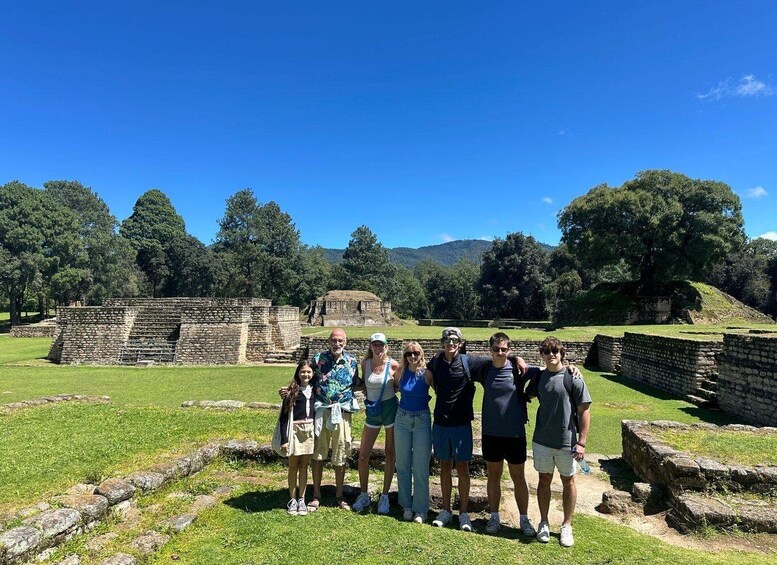 A group of people standing in front of ancient stone ruins with a clear blue sky and green trees in the background. 2