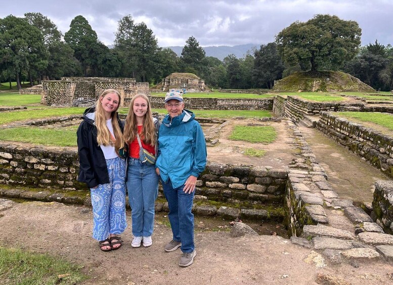A group of people standing in front of ancient stone ruins with a large tree and grassy area in the background. 1
