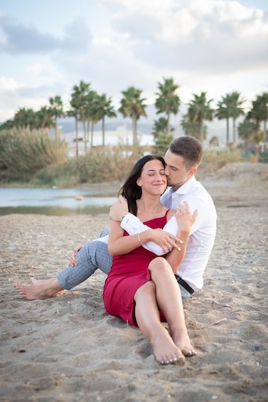 A couple sitting on a sandy beach with palm trees and a body of water in the background. 1