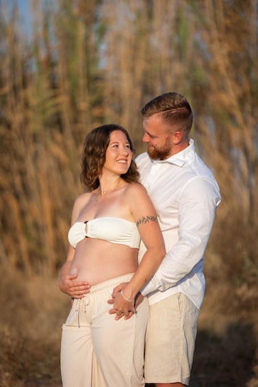 A pregnant woman in a white top and beige pants, holding her belly, with a man in a white shirt and beige shorts standing behind her. 2