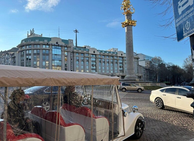 A vintage car with a transparent roof, a golden statue, and a modern building in the background. 2