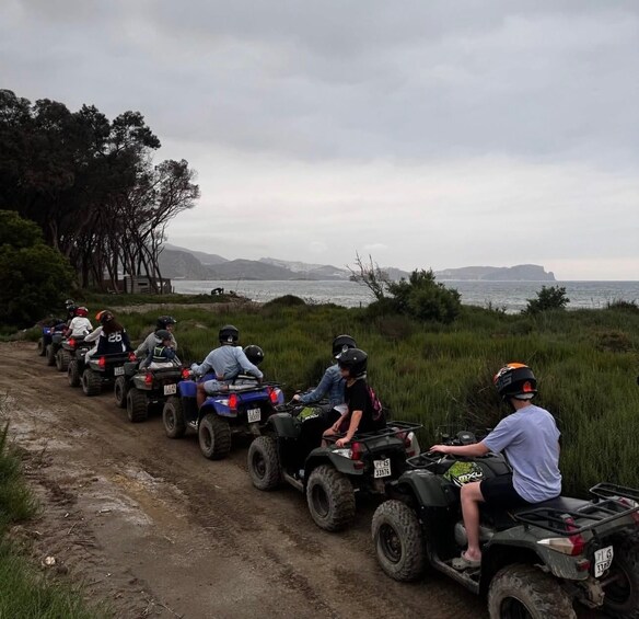 A group of people riding ATVs on a dirt path beside a body of water with trees and hills in the background. 1