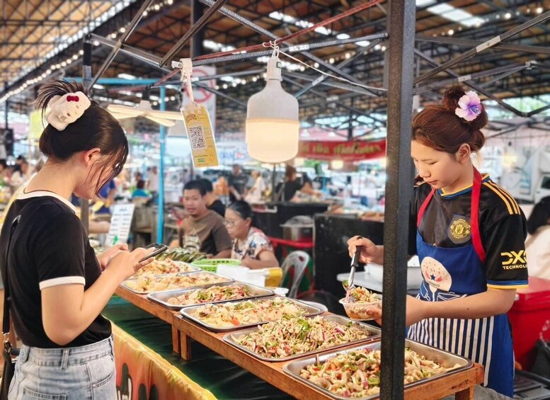 A food stall with various dishes in trays, a hanging light fixture, and a person wearing a blue apron. 5