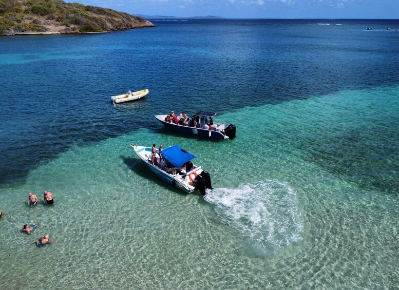 A clear blue sea with three boats, one with a blue canopy, and people swimming in shallow water. 4