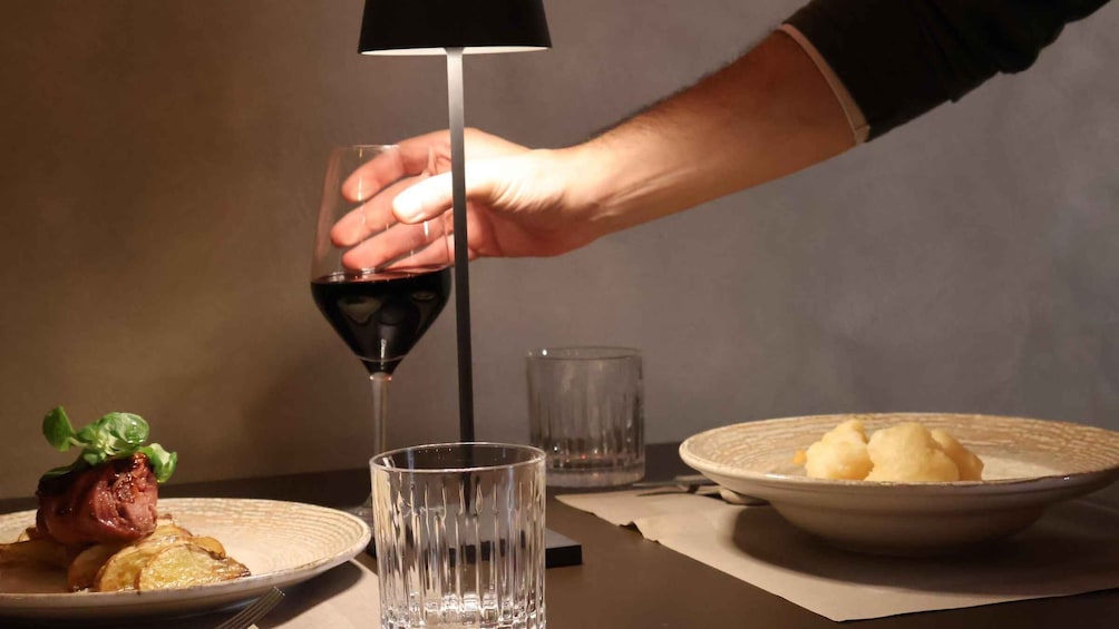 A person pouring red wine into a glass, a plate with food, and a bowl of potatoes. 3