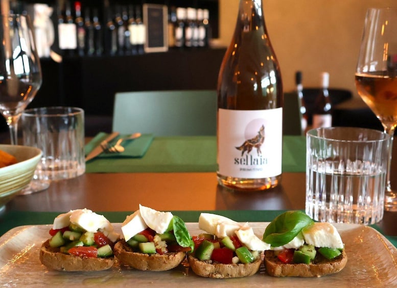 A table with bruschetta topped with vegetables and cheese, a bottle of wine, and glasses. 1