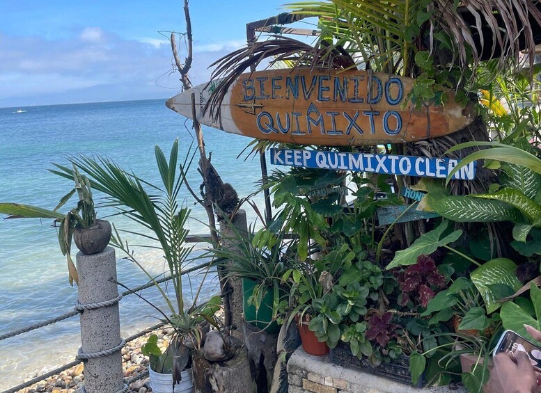 A wooden sign with 'BIENVENIDO QUIMIXTO' and 'KEEP QUIMIXTO CLEAN' written on it, surrounded by tropical plants. 3