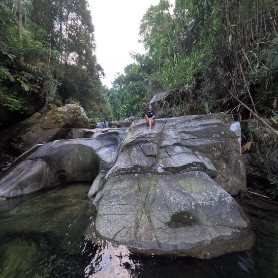 A person sitting on a large rock in a forested area with a stream flowing through. 5