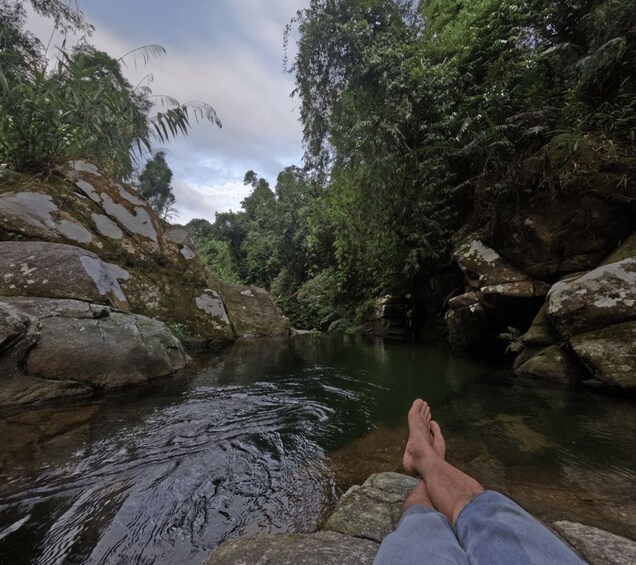 A person sitting by a clear stream surrounded by rocks and lush greenery. 3