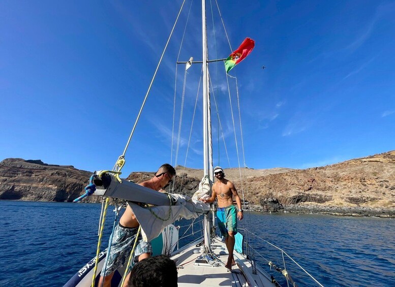 A sailboat with two people on deck, a clear sky, and rocky terrain in the background. 3