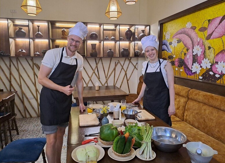 Two chefs in a kitchen with a variety of vegetables, cooking utensils, and a decorative vase wall. 4