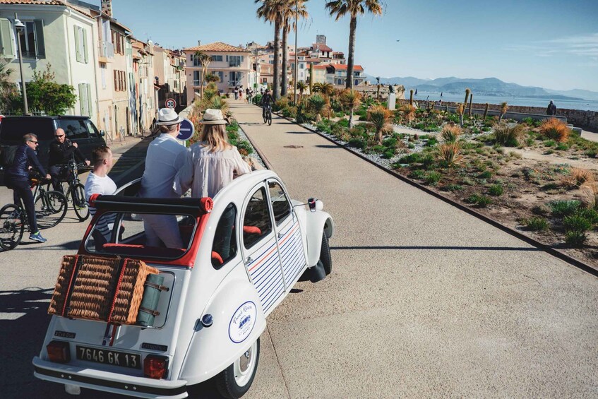 A vintage car with a wicker basket on the back, parked on a coastal road with palm trees and a sea view. 2