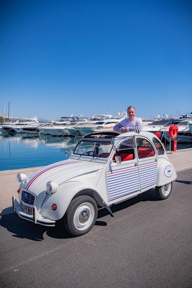 A vintage car with a man sitting in it, parked by a marina with boats in the background. 1