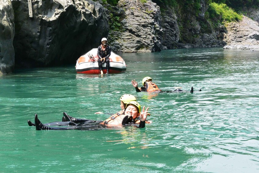 People in a river wearing life jackets and helmets, with a boat and rocky cliffs in the background. 1