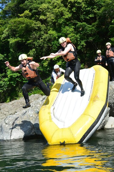People in helmets and life vests on a yellow inflatable raft near rocks and water. 4